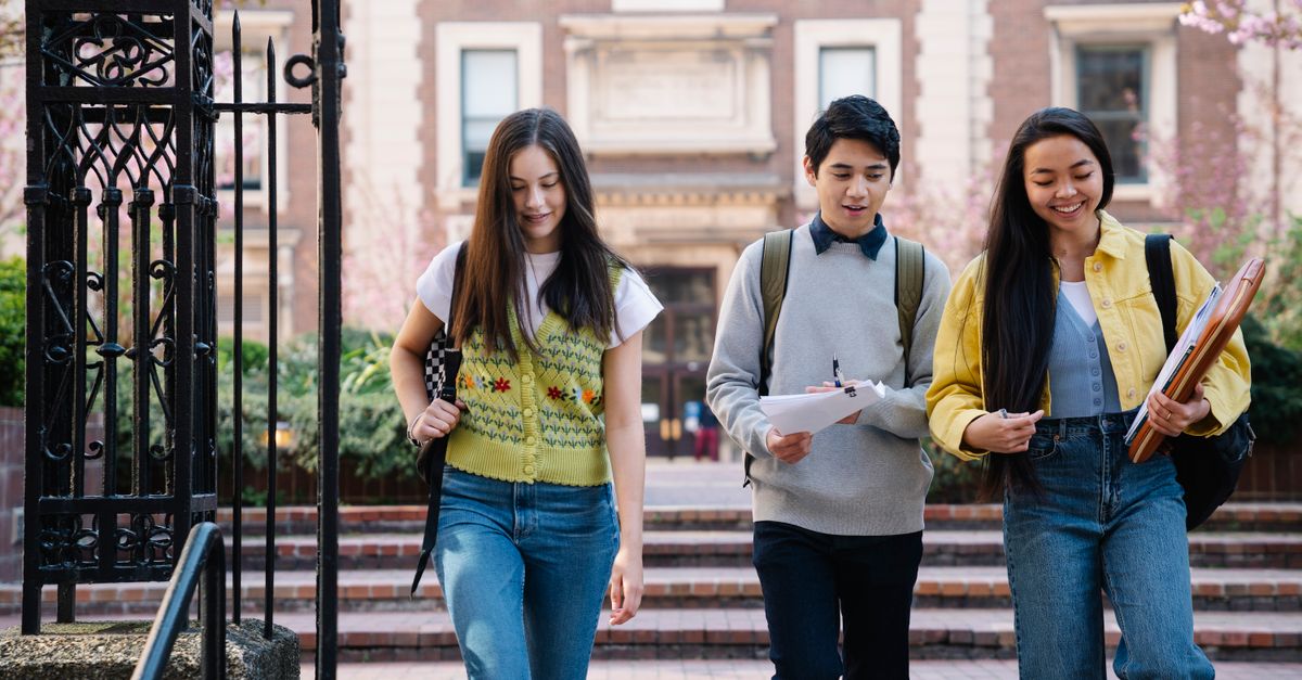 Three smiling students walking together on a university campus, holding notebooks and backpacks.